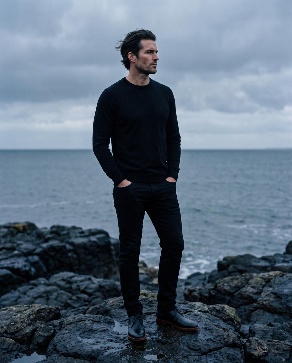 A man in a black knit on gray coastal rocks, wearing the Austin chukka