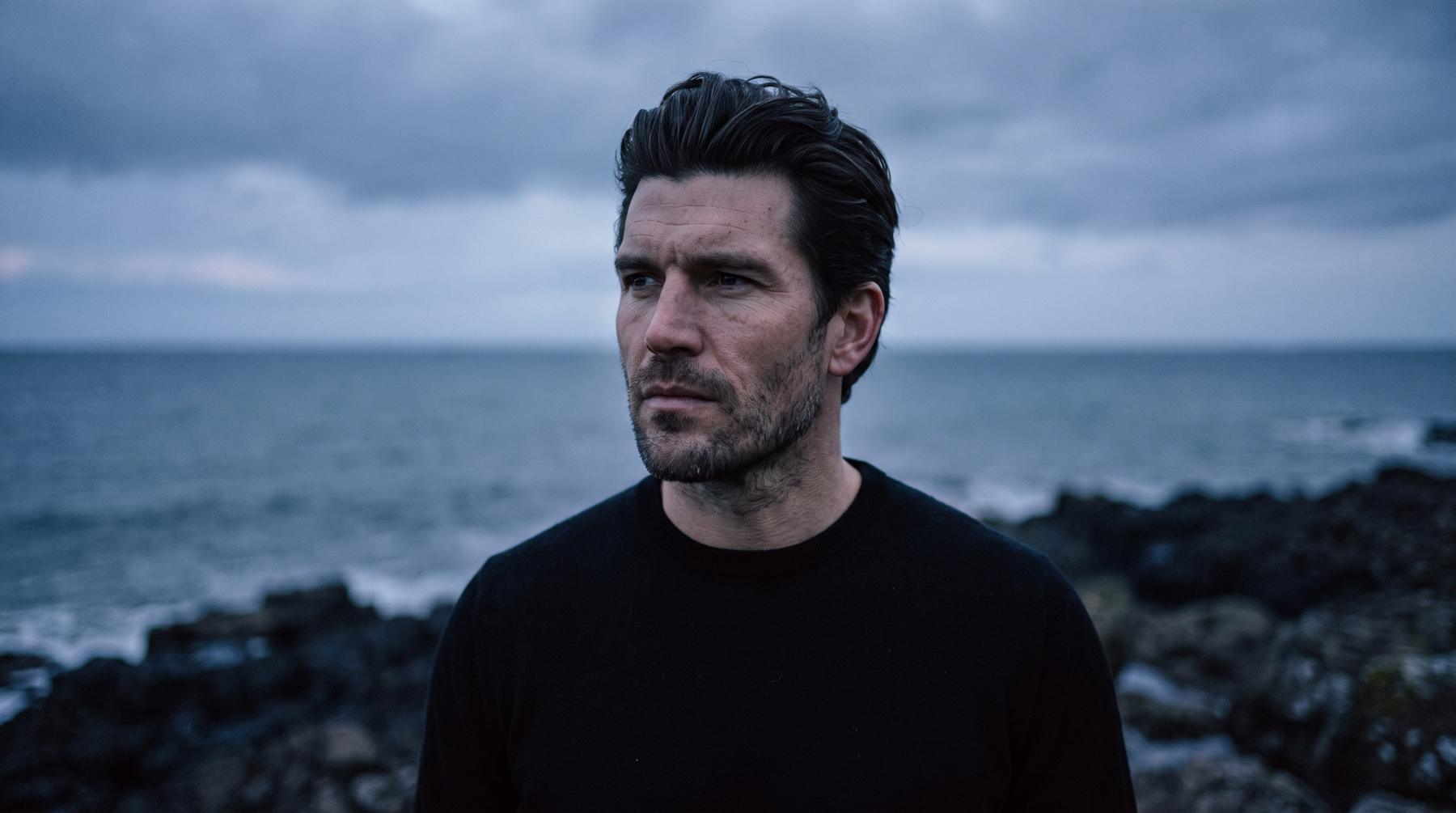 A man in a black crewneck on a rocky shoreline at blue hour